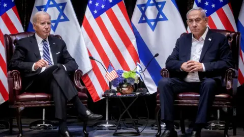 Reuters In front of US and Israeli flags, Joe Biden sits with his legs crossed while to his left sits Israeli Benjamin Netanyahu who clutches his hands in front of his chest