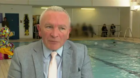 BBC Gary Gentle is sitting in front of a swimming pool, where there are children swimming. He has white hair and is wearing a grey suit jacket, blue shirt and salmon coloured tie.