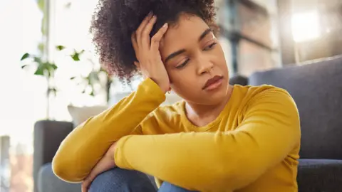 Getty Images Stock image of a woman looking sad and thoughtful, sitting on the floor by a blue sofa with her knees hitched up and her left arm across them. She is holding her head with her right hand. She is wearing a yellow top and blue jeans. 