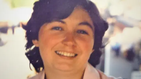 Handout A woman with mid length brown hair smiles towards the camera, the collar of the pink top or coat she is wearing is barely visible. The background is out of focus