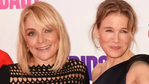 Getty Images Helen Fielding (L) and Renee Zellweger attend the world premiere of Bridget Jones's Baby at Odeon Leicester Square in London in 2016. They both have blonde hair and are wearing black dresses. Both are smiling and looking at the camera.