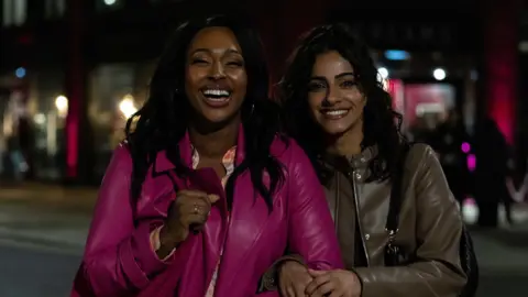 Curfew / Paramount+  Alexandra Burke is wearing a magenta pink jacket and is stood next to Mandip Gill wearing a brown jacket. It is night-time and the two are smiling and laughing at the camera.