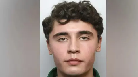 Met Police A mugshot of Daniel Khalife who is looking directly at the lens of a police station camera. His eyes are wide open, he is wearing a green clothing and has black curly hair