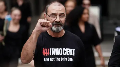 PA Media Andrew Malkinson in a black t-shirt bearing the words '#innocent and not the only one' with his first raised surrounded by other people. 

