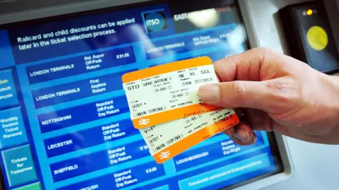 PA Media Two kinds of rail tickets are held up in front of a ticket machine displaying a variety of destinations in the UK