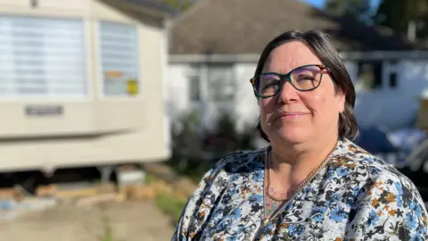 BBC Phoenix Graham stands in the driveway of her home in Ruskington, Lincolnshire. She is wearing a floral blouse and dark-rimmed glasses. In the background stands a cream-coloured static caravan, next to a white-walled bungalow.