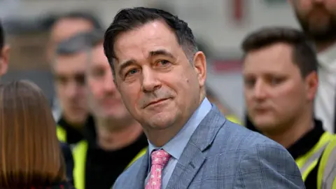 Getty Images GB Energy chairman Juergen Maier stands in front of men in high-vis jackets. He wears a grey suit with a light blue striped shirt and a pink tie with flamingos on it. He has short dark hair.