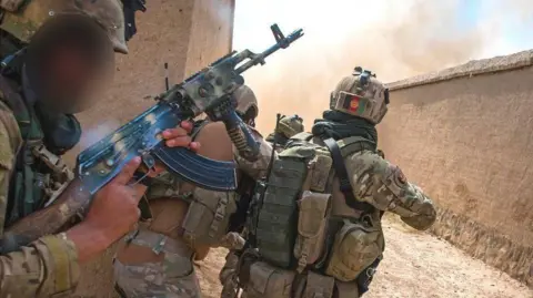 Ben Taggart Three Afghan soldiers in camouflage gear with guns peer around the corner of a sand-coloured building, with smoke seen around the corner