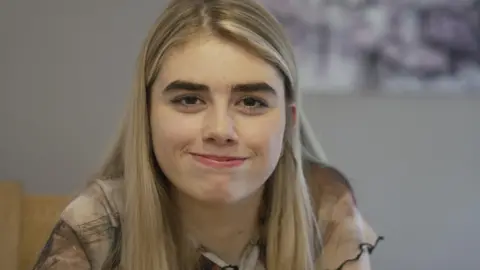 BBC A teenage girl with blonde hair and brown eye looks directly into the camera smiling, the background is out of focus and grey.