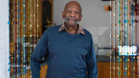 PA Alford Gardner, an elderly gentleman wearing a blue jumper and bushy stubble, smiles into the camera while standing in a kitchen.