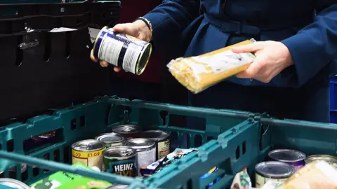 PA Media Close up of a woman's hands as she picks out items from crates at a foodbank. She has a tin in one hand and a packet of spaghetti in the other, with more items in front of her. 