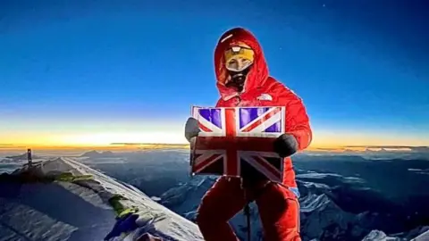 AGA Adventures Adriana Brownlee stands at the peak of a mountain in a red snowsuit, yellow hat and black mask, holding a Union Jack flag, with a sunset behind her