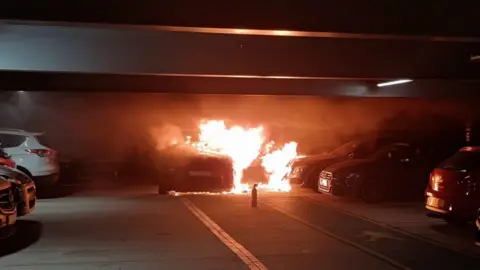 Bedfordshire Fire & Rescue Service A car is on fire inside a multi-storey car park. It is not parked in a bay. There is an abandoned fire extinguisher on the floor. 