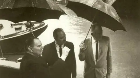 Michael McCulloch A black and white photograph showing Robert McCulloch (right) with his business partner C.V. Wood (centre), standing under umbrellas on a path by the River Thames and looking into the distance