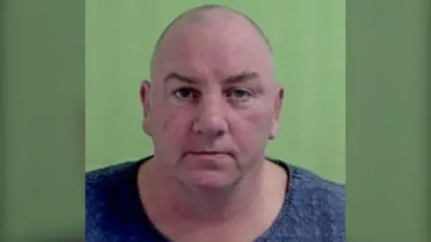 IOM POLICE John Meadows a bald middle-aged man, stares directly at the camera in a police mugshot with a green background.