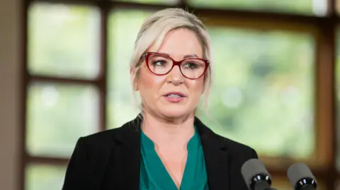 PA Michelle O'Neill in a black blazer and green blouse with red glasses. She has blonde hair tied up behind her head and is standing in front of two black microphones.