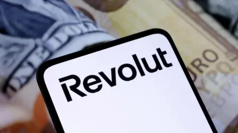 Reuters A photograph of a mobile phone showing the Revolut brand name with bank notes in the background behind the device