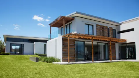 Getty Images A large modern house with whitewashed walls and wooden elements. The house is situated within a grass lawn and under a blue sky with a couple of clouds.