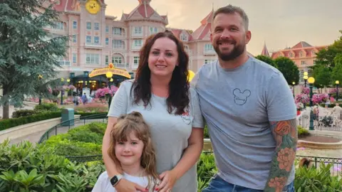 Angharad Dennis Angharad Dennis and husband Joe are wearing grey t-shirts with the Mickey Mouse logo. They stand in a park at Disneyland smiling with their daughter Cari.