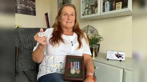 Kevin Shoesmith/BBC Angie Moore, with long brown hair, is pictured holding her son's dog tags and Afghanistan medals