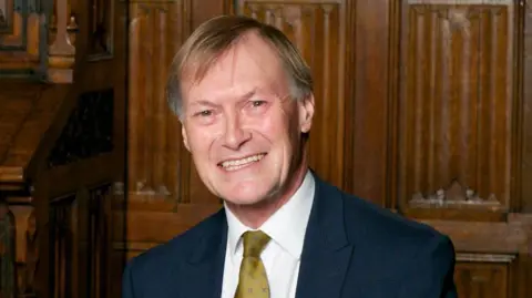 Getty Images Sir David Amess, wearing a dark suit, white shirt and gold-coloured tie, smiles at the camera. He is seated. Behind him is a wood-panelled wall.