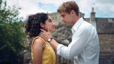 Ludovic Robert/Netflix A young man in a white shirt - Dexter - holds the face of a young woman in a yellow top - Emma. They are staring into each other eyes, and standing in front of a distinctive Edinburgh backdrop. 
