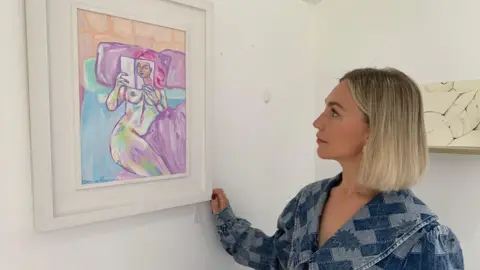 BBC Clíodhna Doherty looks at her painting Laura in her art studio in Newry. The painting is a piece of abstract figurative art showing a naked woman reading a book while in bed. Clíodhna has a blonde bob and is wearing a blue denim dress.