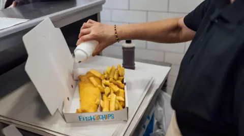 Getty Images A man pouring salt on to a portion of fish and chips in a takeaway box behind the counter of a fish and chip shop