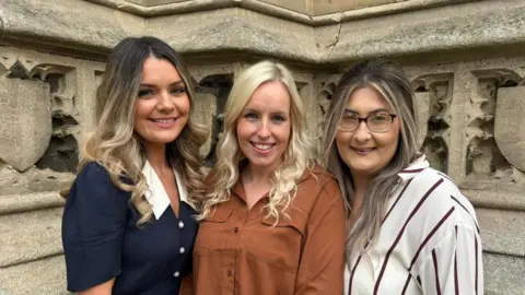 GOSH Louise Hubbard, Justine Ducker and Danielle Mann smiling at the camera. Louise is wearing a blue dress with white buttons and a white collar. Justine is wearing a brown shirt. Danielle is wearing a red and white striped shirt and wears glasses