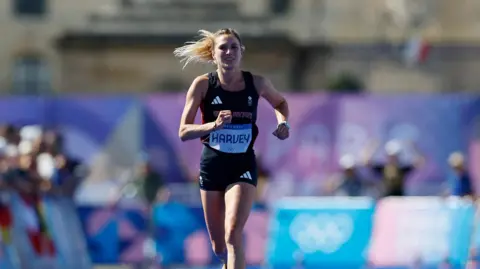 Reuters Rose Harvey in black running kit with her tied-back blonde hair flowing behind her against a blurred background of Olympic branding and supporters. 