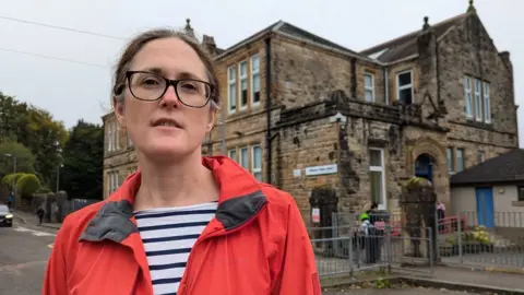 BBC Karen Reid stands in front of Milngavie Primary School. She is wearing a stripy top, and red jacket and glasses with a neutral expression. The primary school is an old sandstone building, slightly dirty with age.