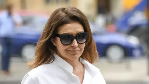 Getty Images Zamira Hajiyeva is wearing sunglasses, a white, buttoned shirt and has brown hair. A blue car, which is out of focus, is behind her