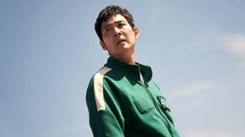 No Ju-han/Netflix Lee Jung-jae as Gi-hun, wearing a green tracksuit against a blue background