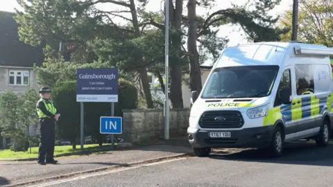 BBC A female police officer stands at the entrance to Gainsborough Care Home. A marked police van is parked on the roadside. The two-storey building can just be seen behind some trees and a blue sign saying 'Gainsborough Care Home'.