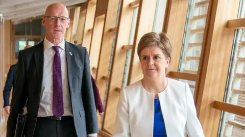 PA Media John Swinney and Nicola Sturgeon waslking down a long corridor at Holyrood. Swinney is on the left of frame in a dark suit and purple tie, carrying a folder. Sturgeon is on the right weating a white collarless jacket and electric blue blouse. 