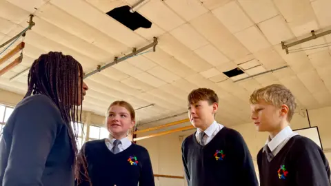 BBC/ Hazel Shearing Four students stand in the gym at Patchway Community School in Bristol. Above them, two ceiling panels are missing and cracks have appeared in others