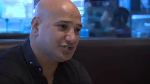 BBC Shabir Hussain, a man with brown eyes, shaven head and wearing a black collared shirt speaks to someone off-camera.