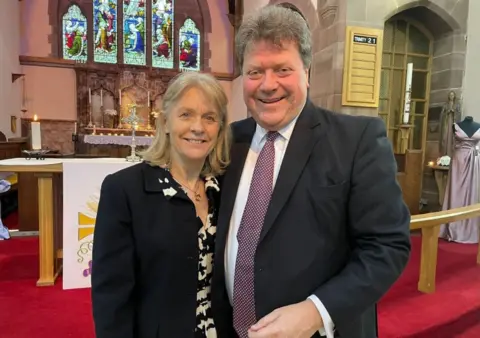 BBC Sue and Alex Tatham inside the church with a stained glass window and candles behind them. Both are wearing jackets and standing on red carpet.