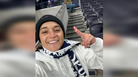 Mandy Damari  Emily Damari, a young woman wearing a Spurs scarf and a black beanie hat, smiles in the stands of a football ground