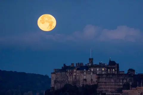 Jane Barlow/PA Media The full October moon, known as the Hunter's sets behind Edinburgh Castle, Scotland. 17 October  2024.   The moon is on the left hand side of a blue dusk sky. The castle in in the foreground to the right.