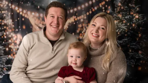 Jo Bishop Photography/PA Media A smiling Sam and Laura hold their one-year-old daughter Aurora in a family photograph shot against Christmas trees covered in fairy lights. 