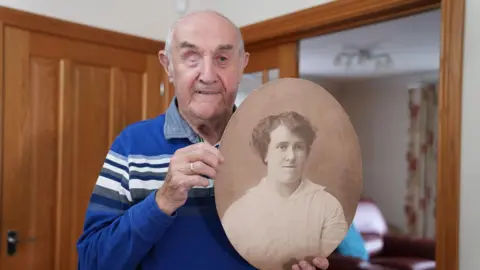 Eddie Wulff holds a large sepia photograph of his grandmother, Mabel Wulff. Dressed in a blue and white stripy polo shirt, Eddie smiles for the camera in his family house. His grandmother's photograph, in his hands, is an old portrait. She wears a white shirt. 