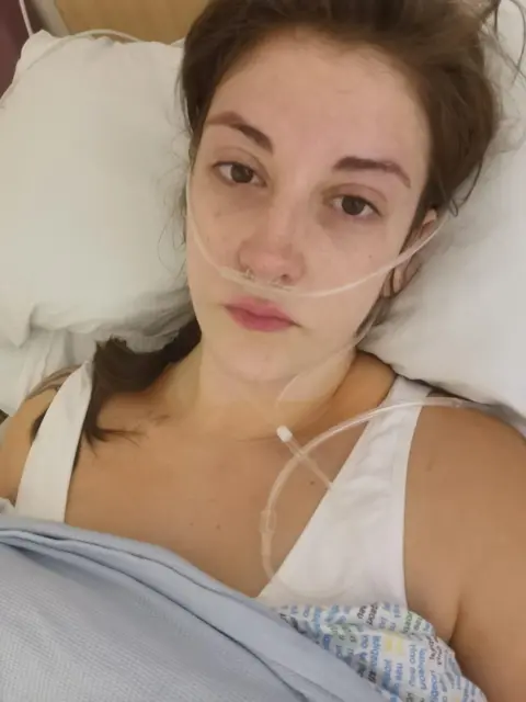 Lisa Bancroft Woman in a hospital bed with an oxygen tube attached to her nose. She has her head on a white pillow and blue sheets pulled over her chest. She is looking at the camera.