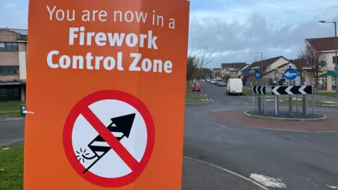 An orange sign with white writing on it reading "you are now in a fireworks control zone". Below is a symbol showing a lit firework on a white background with a red line through it.