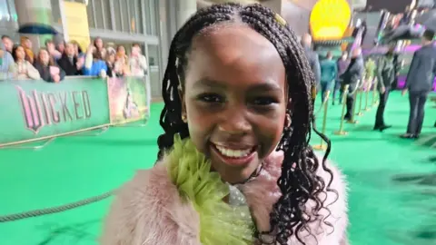 Lauren Kelly A young girl is stood on a green carpet whilst smiling at the camera. She is wearing a green dress and a pink fluffy coat.