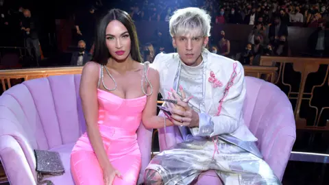 Getty Images Megan Fox, wearing a pink dress, with silver straps and pink fingernails, holding hands with Machine Gun Kelly, wearing Silver shorts and a silver jacket. The pair are sitting on a purple sofa.