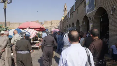 BBC correspondent in a market in Erbil, Iraq. He has his back to the camera and is wearing a light blue shirt with a bag on his right shoulder. There arae several men walking about the street.