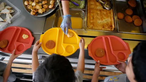 Reuters Three young children photographed from above hold out brightly coloured plastic trays with some food on them as more food is placed on one of the trays. In front of the children is an array of food, including roast potatoes and a pie in a metal tray. 