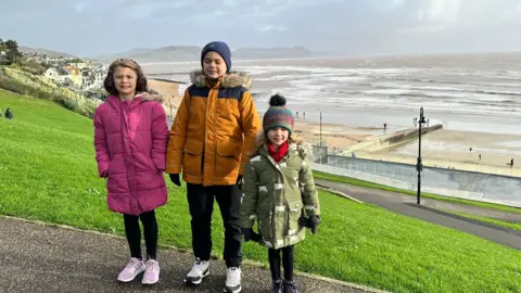 Noah, wearing an orange and black jacket, black trousers and white trainers, is flanked by his two sisters. They are photographed on a coastal path with a beach and the sea below them.