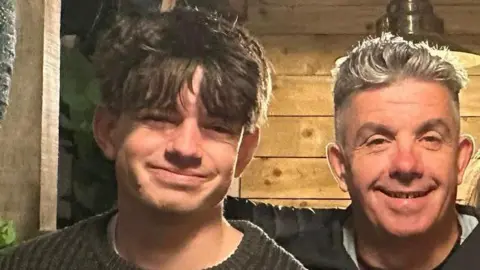 Handout A cropped shot of Sam and his dad smiling at the camera. Sam is wearing a crew-neck knitted jumper and has medium-length brown hair which comes down over his eyebrows. David is wearing a dark coat and has spiky grey hair.